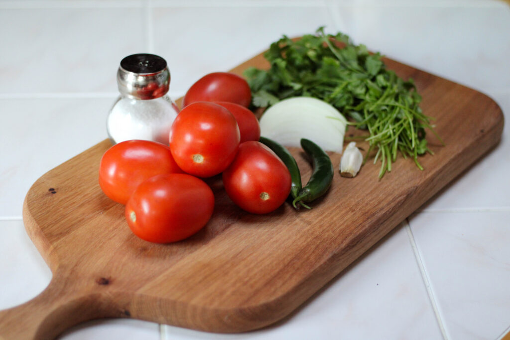 Salsa ingredients on a cutting board with white background including tomato, serrano chilis, garlic, onion, cilantro and salt. Salsa de Molcajete Ingredientes