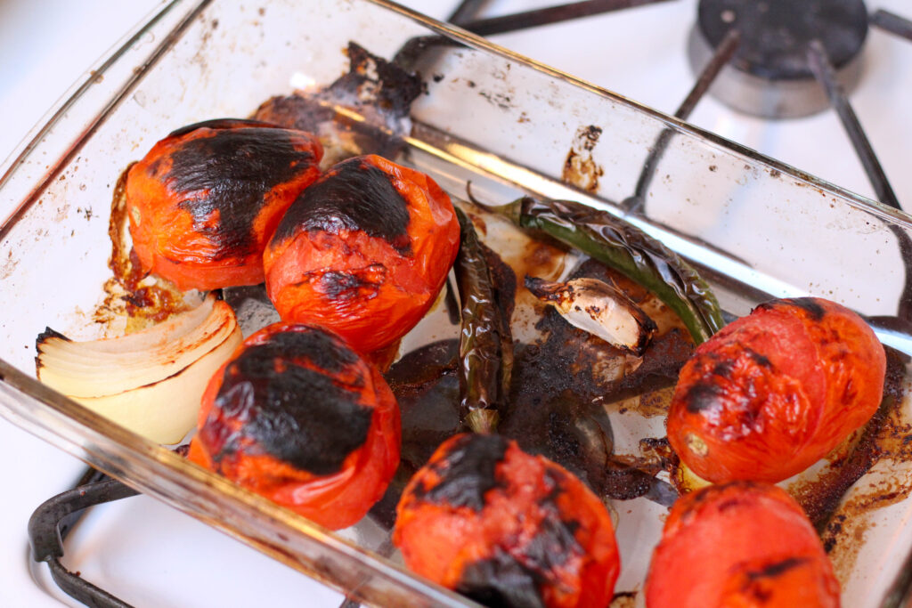 Roasted tomatoes, serrano peppers, onion and garlic in a pyrex dish on top of a stove. 