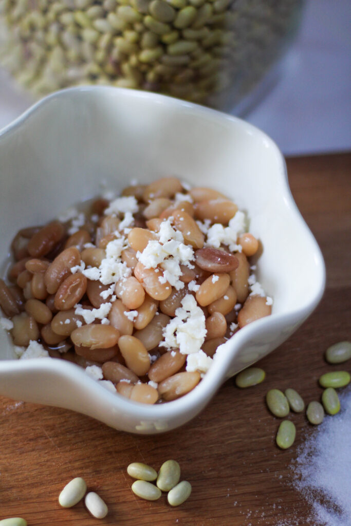 Cooked Peruvian Beans in a white bowl with cheese on top. Peruano Beans topped with crumbled cheese. Mayocoba beans with queso fresco with a cutting board and beans in the background. 