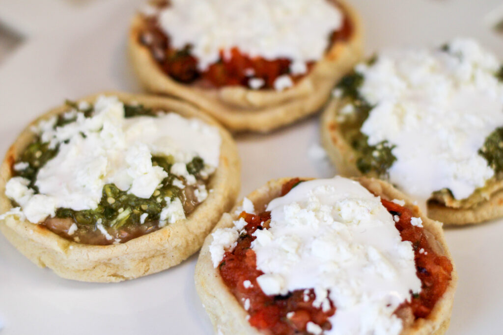 Picaditas topped with beans, salsa, crema and cheese served on a white plate. 