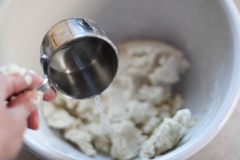 Hand pouring water into bowl with dough using metal measuring cup.