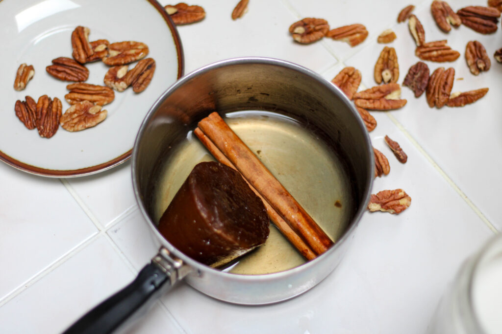 Small saucepan with piloncillo, water and a cinnamon stick on a white tile table with pecans in the background. 