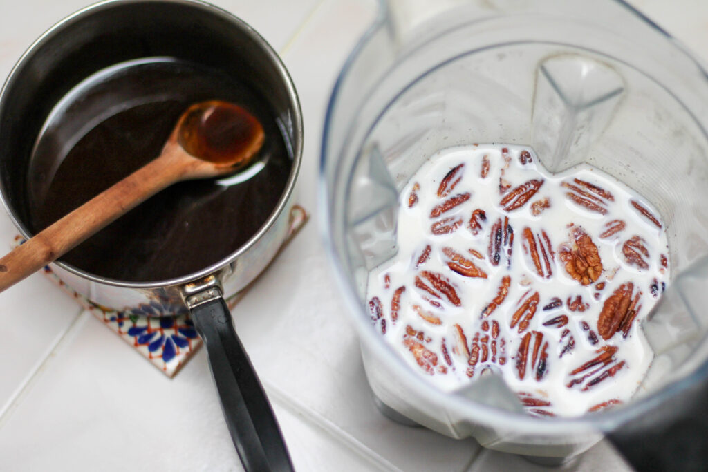 Blender with milk and pecans and a saucepan with dark sauce and wooden spoon. 