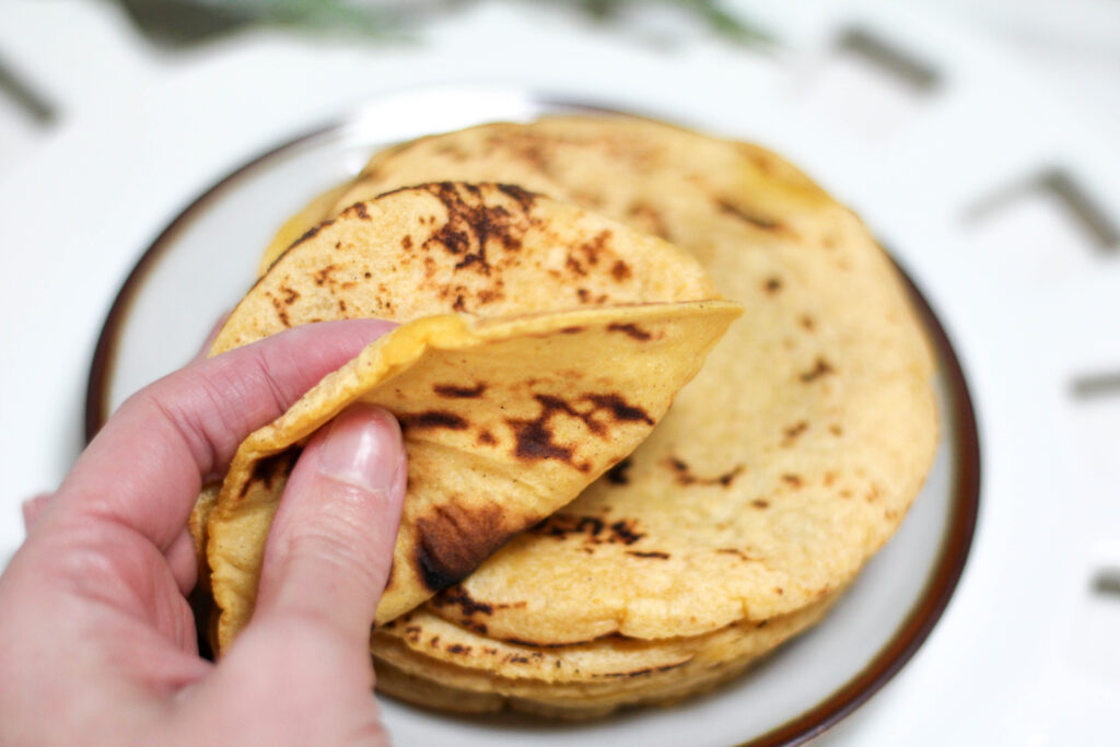 Hand holding handmade corn tortillas stacked on a plate on a white table. 