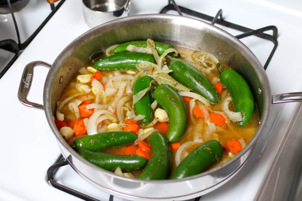 Jalapenos, onion, carrots and spices cooking in a saucepan on a white stove.
