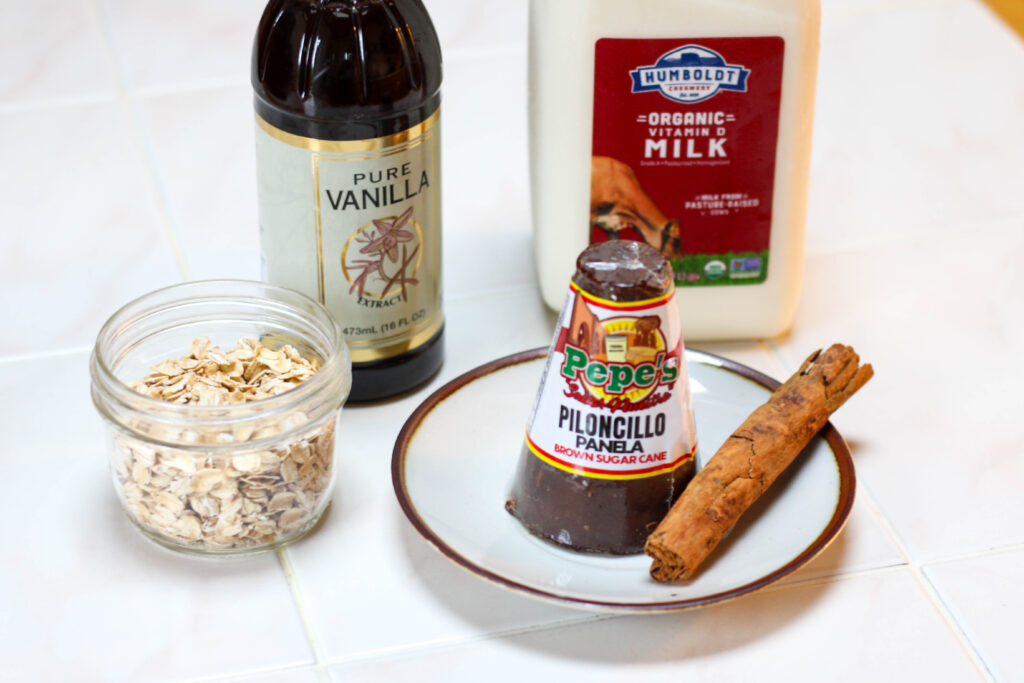 ingredients for atole de avena on a white table including milk, piloncillo, cinnamon stick, oatmeal and vanilla
