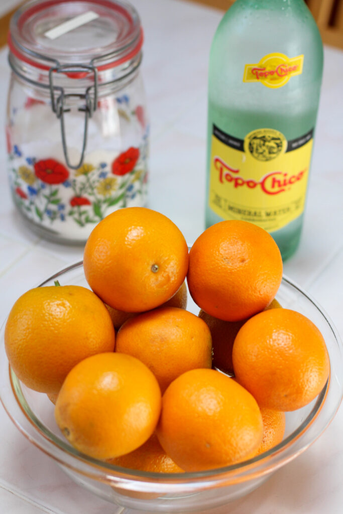 Glass bowl of oranges with jar of sugar and bottle of Topo Chico in the background
