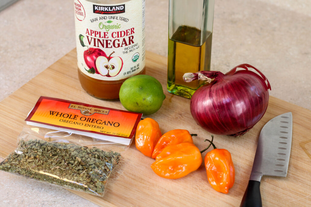 ingredients for cebolla con habanero on a wood cutting board including red onion, habaneros, lime, oregano and apple cider vinegar
