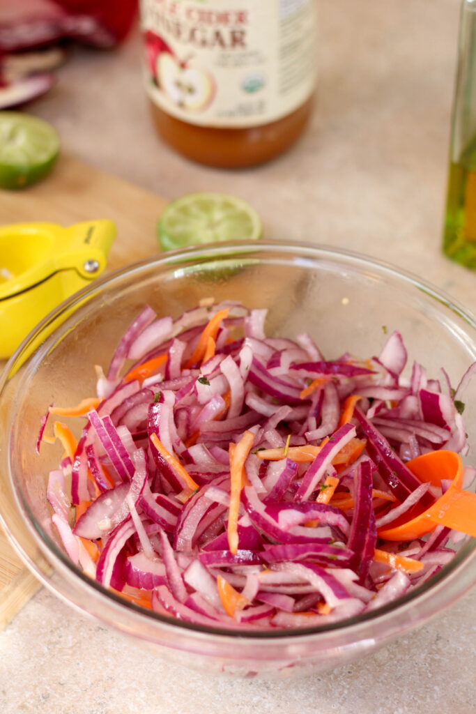 Glass bowl with sliced red onion and sliced habanero on a kitchen counter with food in the background