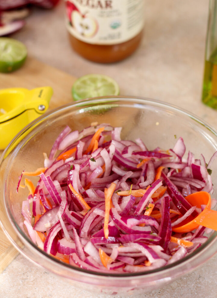 Glass bowl with sliced red onion and sliced habanero on a kitchen counter with food in the background