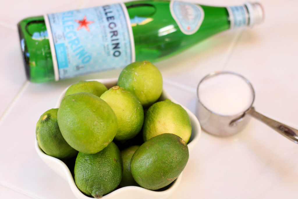 Bowl of limes, cup of sugar an sanpellegrino on a white table
