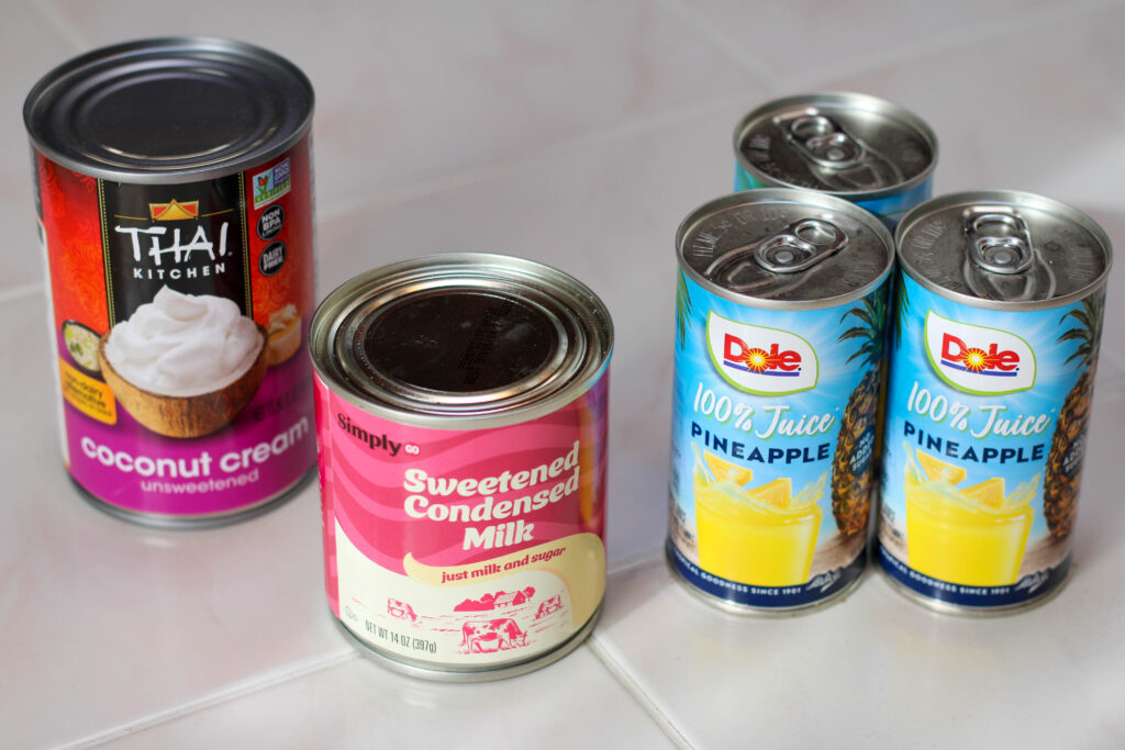 Can of coconut cream, sweetened condensed milk and three cans of dole pineapple juice with white background
