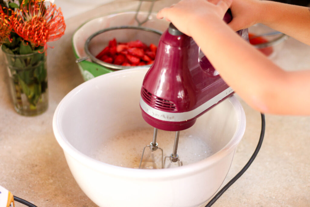 Hands holding purple hand mixer mixing cream and sugar in a white bowl