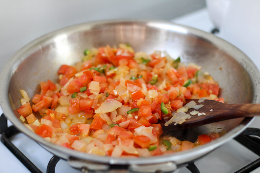 Onion, tomato, garlic and serrano pepper cooking in a stainless steel saute pan on a white stove