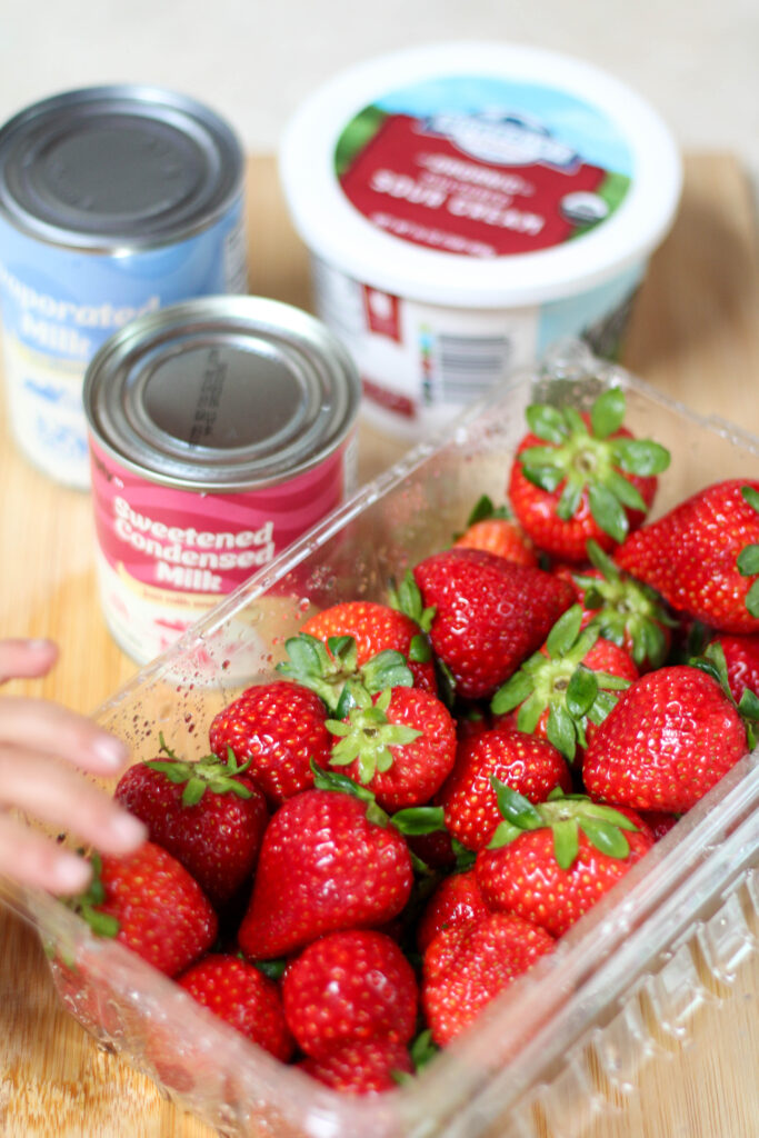 Strawberries, sour cream, sweetened condensed milk and evaporated milk on a wood cutting board