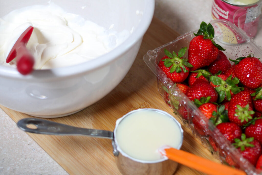 White bowl with cream, cup with sweetened condensed milk and strawberries on a wood cutting board