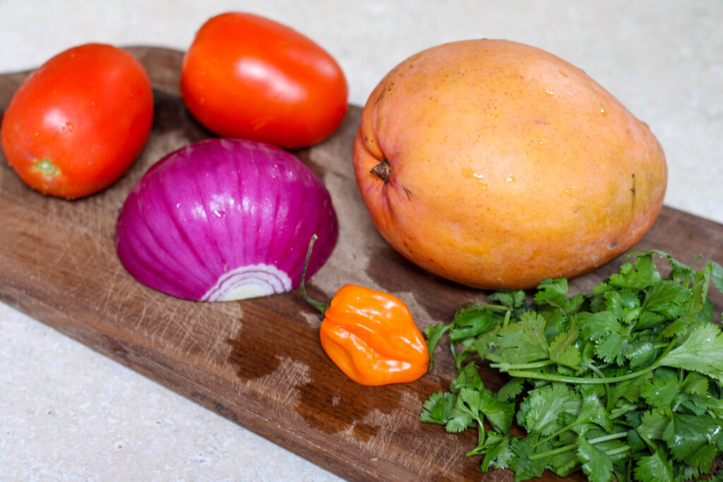 One mango, two tomatoes, red onion, habanero pepper and cilantro on a wood cutting board