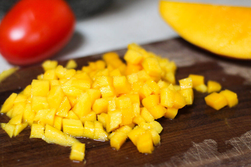 Diced mango on wood cutting board with red tomato in the background